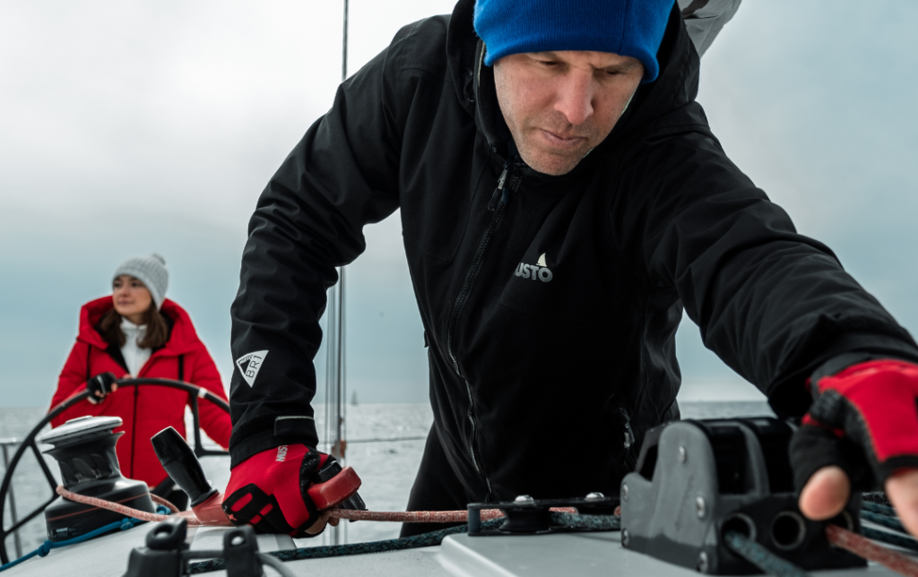 Two sailors dressed for winter sailing in the Bay, focusing on handling ropes and steering under cool, calm conditions.